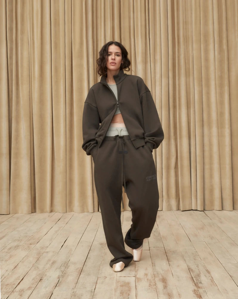 นางแบบใส่ชุดวอร์มโทนเข้มจาก Fear of God ESSENTIALS ลุคเรียบหรูแบบ Quiet Luxury ผสมกลิ่นอายสตรีทแฟชั่น ถ่ายภาพหน้าฉากผ้าม่านพื้นไม้ในสตูดิโอ