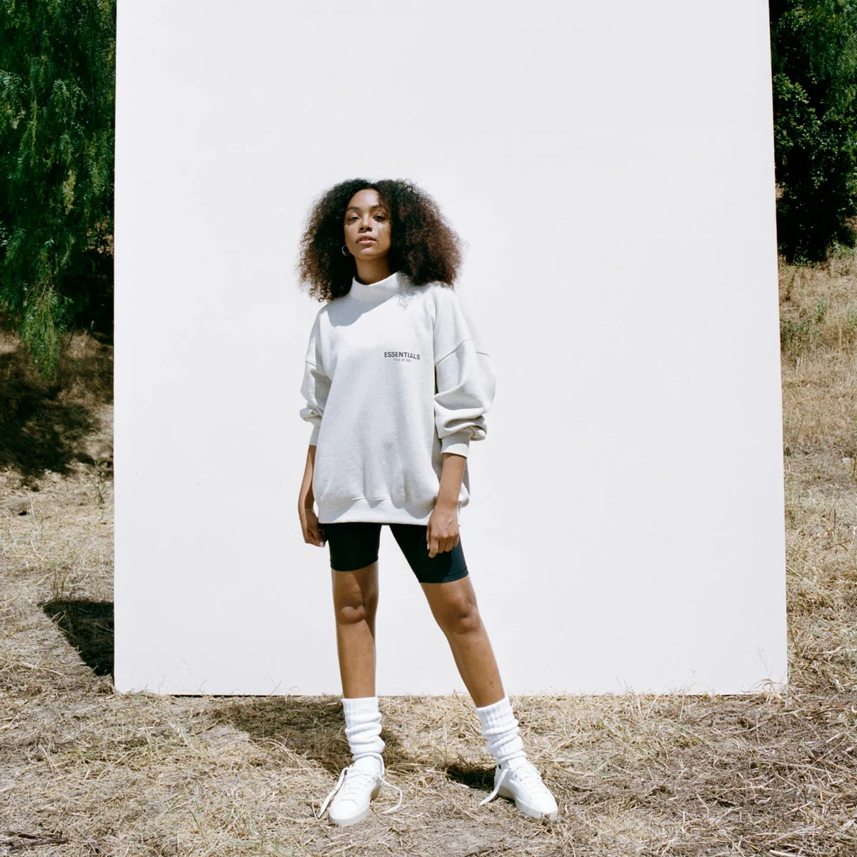 นางแบบสวมเสื้อสเวตเตอร์ Fear of God ESSENTIALS สีครีมยืนหน้าพื้นหลังสีขาวท่ามกลางธรรมชาติ ลุคมินิมอลแนว streetwear โทนเรียบหรูแบบ Quiet Luxury