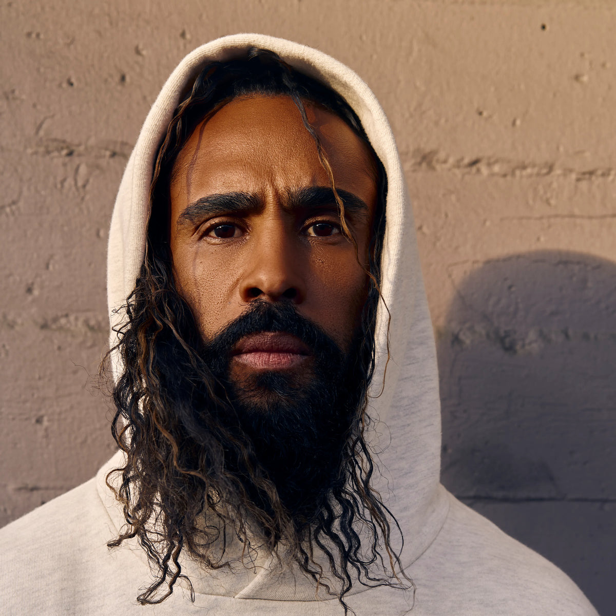 Jerry Lorenzo ผู้ก่อตั้งแบรนด์ Fear of God ESSENTIALS ใส่เสื้อฮู้ดสีครีม ยืนหน้าผนังคอนกรีตในแสงธรรมชาติ โทนภาพเรียบหรู สื่อถึงสไตล์ Quiet Luxury แบบดิบเท่